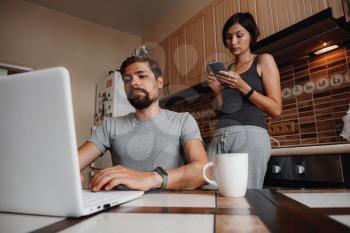Young couple using laptop and smartphone in the kitchen, smiling man working online at home, family morning lifestyle with gadgets or devices addiction concept. readings news
