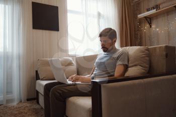 Pensive bearded man sitting on sofa work at laptop thinking of problem solution, thoughtful male employee pondering considering idea looking at computer screen making decision