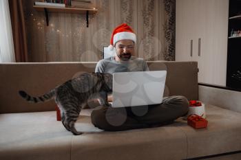 A hipster man with grey cat sitting on a sofa at home at Christmas time. Virtual Christmas house party. Online team meeting video conference calling from home. Man wearing Santa hat