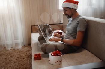 A hipster man with grey cat sitting on a sofa at home at Christmas time. Virtual Christmas house party. Online team meeting video conference calling from home. Man wearing Santa hat