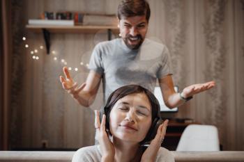 angry husband shouting at wife sitting on sofa and covering ears with headphones. the woman ignores the man