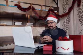 Happy male in deep blue shirt makes a purchase on the Internet via a phone. Lots of scenery for the new year. Working from home businessman wearing red Santa hat