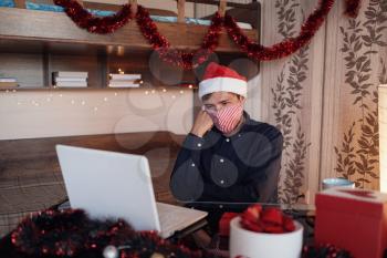 portrait of unhappy man in mask sitting in room near christmas decorations with lights and ordering gifts. New Year e-shopping. Merry Christmas Covid 19 coronavirus social distance concept.