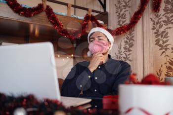 portrait of unhappy man in mask sitting in room near christmas decorations with lights and ordering gifts. New Year e-shopping. Merry Christmas Covid 19 coronavirus social distance concept.