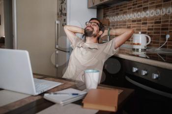 Smiling hipster freelancer holding hands behind head sitting at office desk behind laptop. Happy employee feeling no stress, relaxing, watching funny video after successful working