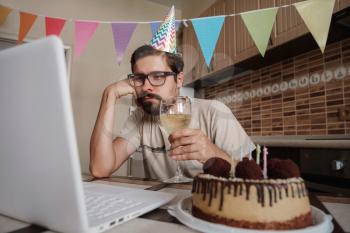 Upset young man sitting at the birthday cake and looking with sad eyes on it. Concept of solitude in quarantine during the Coronavirus Pandemic COVID-19.