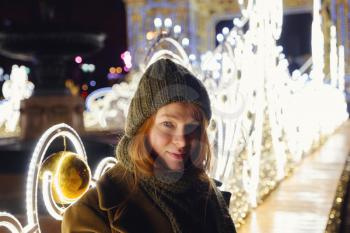 young smiling woman posing on the street. Festive Christmas fair in the background. Model wearing a stylish winter coat, knitted cap, scarf. Feeling happy in big city. Spending winter vacations Moscow