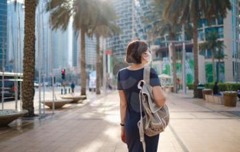 Portrait of young woman wearing in blue dress and white mask for prevent virus, walk in front of skycrapers in modern city. view from the back or rear view, the lady walks away from the photo camera