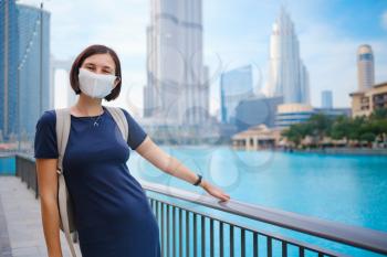 Portrait of beautiful woman wearing a mask for prevent virus walking in Dubai with skyscrapers in the background. Enjoying travel in United Arabian Emirates. Waiting for the evening fountain show