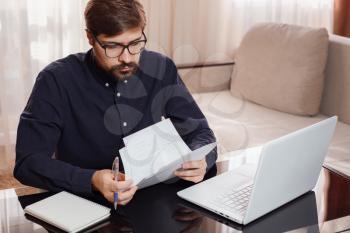 Entrepreneur in eyeglasses works with a laptop and keeps a document in a home office. Man holding paper documents, chatting online with clients on laptop at workplace.