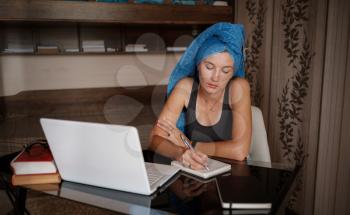 Woman with blue towel on head is working office work remotely from home. Using computer. Distance learning online education and work. Freelancer working