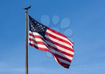 Wind blows the USA flag of stars and stripes with eagle flagpole against blue sky late in the afternoon as sun sets