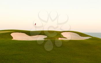Red flag of golf hole above sand trap or bunker on beautiful ocean front course at sunset