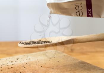 Superfood known as black chia seeds are poured from packet into a wooden backing spoon over wood bread board on kitchen table