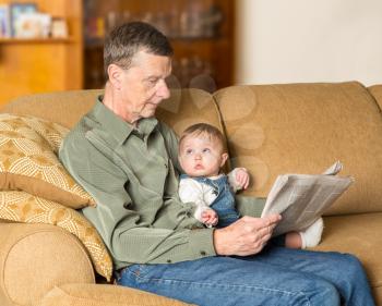Young caucasian baby girl looking up at grandfather reading newspaper on settee in family room
