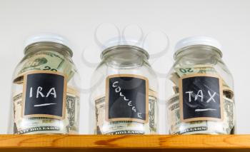 Three glass jars with chalk labels used for saving US dollar bills and notes for IRA, tax and college funds