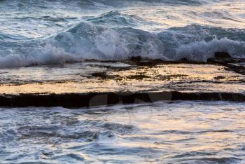 Water pool on rock shelf reflects the orange sky at sunrise as ocean waves crash at Moloaa Beach, Kauai, Hawaii
