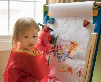 Young two year old girl drawing with crayon on paper mounted on an easel during playtime