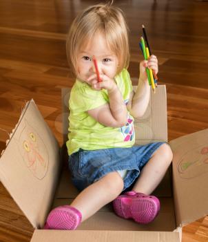 Young two year old girl sitting in cardboard box and decorating it with crayons and coloring