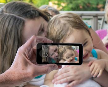 Woman and small baby girl laughing and cuddling in photo being taken by male hand on smartphone