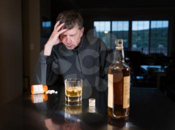 Senior adult male facing a kitchen table with alcoholic drink and looking very sad and depressed with tablets or pills