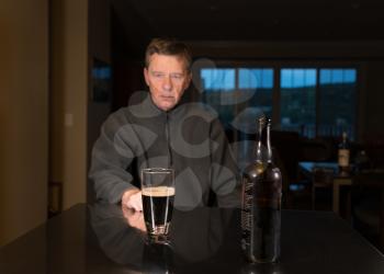 Senior adult male facing a kitchen table with alcoholic drink and looking very sad and depressed