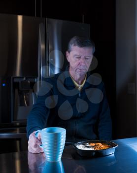 Lonely and depressed senior male sitting alone at kitchen table eating a microwaved ready meal of curry and mug of tea or coffee