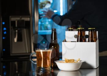 Six pack of brown beer bottles in plain white cardboard carrier with mug of ale on stainless steel kitchen or bar counter. Open fridge or refrigerator out of focus in rear.