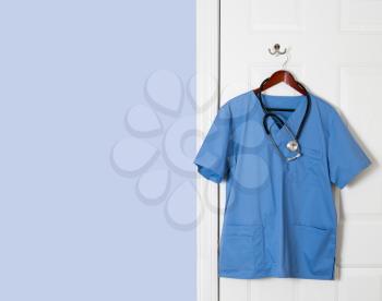 Blue medical scrubs uniform shirt hanging on a hook on back of door with stethoscope with copy space for message
