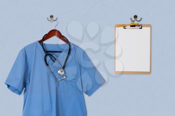 Blue medical scrubs uniform shirt hanging on a hanger with stethoscope and on hanger and hook on blue wall with clipboard for message