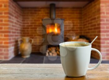 Coffee cup on wooden table in front of roaring fire inside wood burning stove in brick fireplace