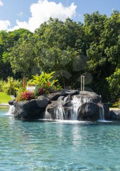 Hot tropical swimming pool at resort with waterfall feature and reclining chairs on the patio with turquiose water