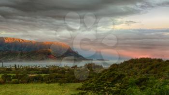 Bay at Hanalei in Kauai with the Na Pali mountain range in the background. Taken just after dawn with the rising sun skimming the peaks of the mountains and casting a sunbeam ray into the ocean