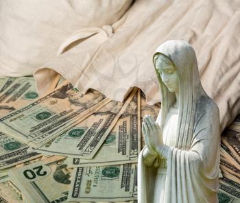 Image of statue of lady praying with US currency as background as a concept for the linkage of religion with money and wealth