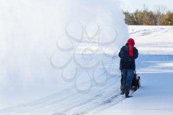 Senior lady using a snowblower on rural drive on windy day with a cloud or blizzard of snow blowing in the air
