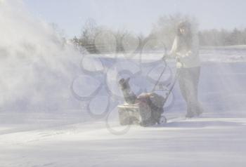 Young lady using a snowblower on rural drive on windy day with a cloud or blizzard of snow blowing in the air and covering her with icy snow