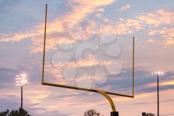 Yellow american football goal posts at school field against the lights and clouds of setting sun