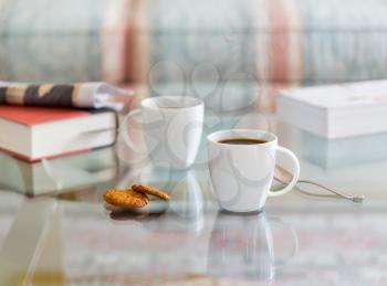 Modern white porcelain cup of black coffee on glass table with spoon and ginger biscuits. Book newspaper in the background out of focus