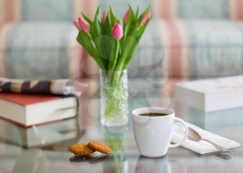 Modern white porcelain cup of black coffee on glass table with spoon and ginger biscuits. Book newspaper and vase of tulips in the background out of focus