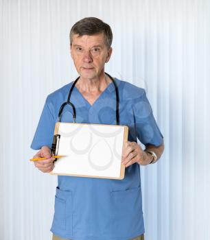 Senior male caucasian doctor with stethoscope in medical scrubs and confidently holding clipboard for message