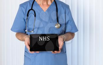 Senior male caucasian doctor with stethoscope in medical scrubs looking up and holding electronic tablet for NHS message
