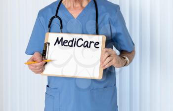 Senior male caucasian doctor with stethoscope in medical scrubs and holding clipboard for Medicare message with pencil for emphasis