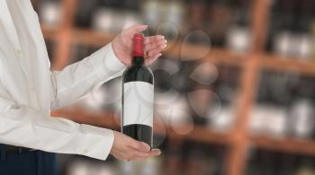 Senior female caucasian wine steward offering a bottle of red wine with blank label to a customer in a wine store or winery. The bottle is ready with copy space.