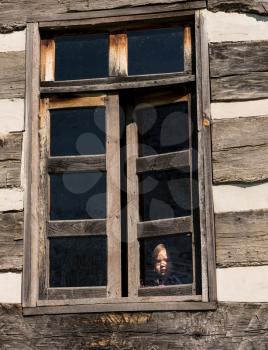 Small and lonely caucasian baby girl or toddler looking out of old wooden cabin window to suggest poverty or recession