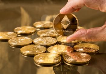 Hand holding a single ether or ethereum coin over bitcoins on gold background to illustrate blockchain and cyber currency