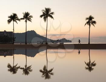 Sunrise in Waikiki with Diamond Head reflected in calm waters of pond as walkers walk along the beach