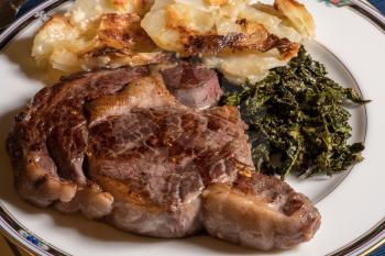 Large grilled ribeye steak on white plate with roasted kale and sliced potatoes