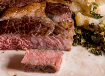 Large grilled ribeye steak with slice on white plate with roasted kale and sliced potatoes
