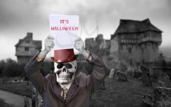 Man wearing suit with a skull and red hat and holding up a message about halloween in a graveyard