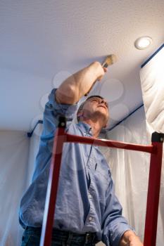 Senior adult male on ladder painting the ceiling of a room inside his home